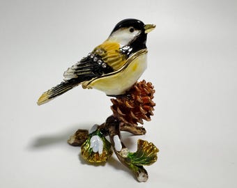 Op de afbeelding: Decoratief vogelbeeldje dat op een tak met een dennenappel zit. De vogel heeft een zwarte kop, een wit en geel lichaam en strass accenten. De tak heeft groene en gouden bladeren.