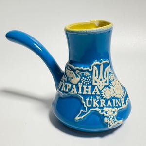 Op de afbeelding: Een blauwe keramische koffiepot met een lange handgreep en een gele rand. De pot heeft een witte omtrek van het land Oekraïne, met het woord "UKRAINE" en het Oekraïense wapen.