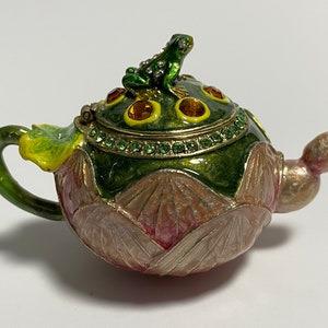 May include: A green and pink teapot with a frog on the lid. The teapot is decorated with rhinestones and has a floral design.