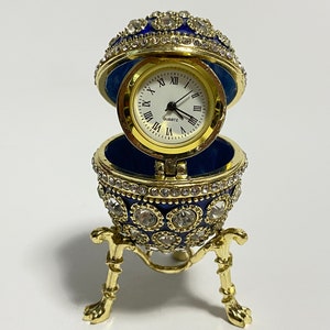 Puede incluir: Un reloj dorado en forma de huevo, enjoyado, con interior azul. La esfera del reloj es blanca con números romanos negros. El huevo está abierto, revelando el reloj, y se apoya en un soporte dorado con tres patas.