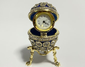 Könnte beinhalten: Eine goldfarbene, mit Juwelen besetzte eiförmige Uhr mit blauem Innenraum. Das Zifferblatt ist weiß mit schwarzen römischen Ziffern. Das Ei ist geöffnet und zeigt die Uhr, die auf einem goldfarbenen Ständer mit drei Beinen steht.