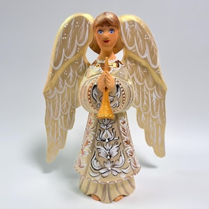 May include: A hand-painted angel figurine with a trumpet. The angel has blonde hair, blue eyes, and a serene expression. The wings are gold with white accents. The angel's dress is cream-colored with floral patterns.