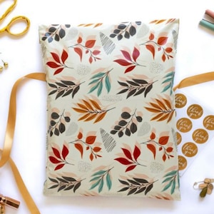 May include: A cream-colored padded mailer with a repeating pattern of colorful leaves in red, orange, teal, and black. Gold scissors, ribbon, and "thank you" stickers are arranged around the mailer, suggesting packaging supplies.