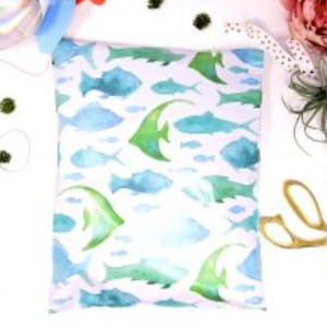 May include: A rectangular, white fabric pouch with a watercolor pattern of blue and green fish. The fish vary in size and shape, creating an aquatic theme. The pouch is likely used for storage or packaging.