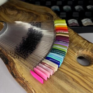 Könnte beinhalten: Ein fächerförmiger Nagellack-Farbkarte mit einer Vielzahl von Farben, darunter Pink, Blau, Grün und Braun. Die Karte ist aus klarem Kunststoff und hat einen Holzgriff.