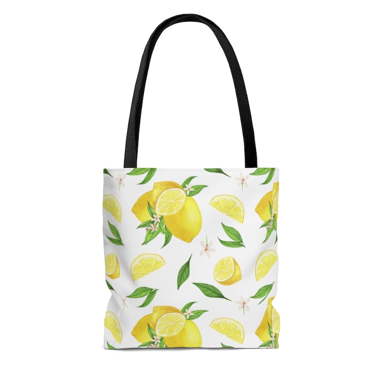 May include: A white tote bag with black handles features a repeating pattern of yellow lemons, green leaves, and white flowers.