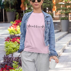May include: A woman wearing a pink t-shirt with a black graphic of a heartbeat with the word "coffee" inside. She is wearing a denim jacket and grey sweatpants. She is holding a white to-go cup.