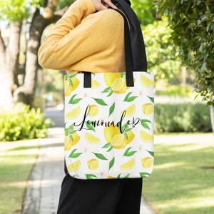 May include: A white tote bag with black handles and a lemon print. The bag has the word "Lemonade" written in black cursive script.