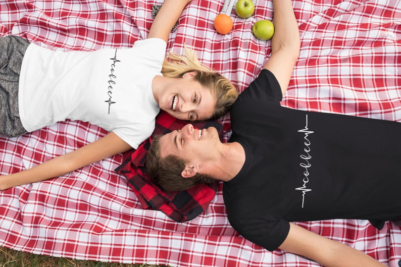 May include: A couple wearing matching t-shirts with a heartbeat graphic and the text "woo-hoo-eep" on a red and white checkered blanket.