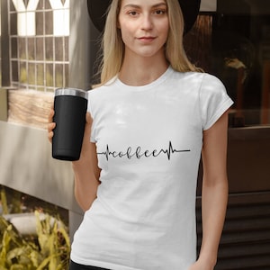 May include: A white t-shirt with a black heartbeat graphic that says "coffee" inside the heartbeat. A person is wearing the shirt and holding a black travel mug.