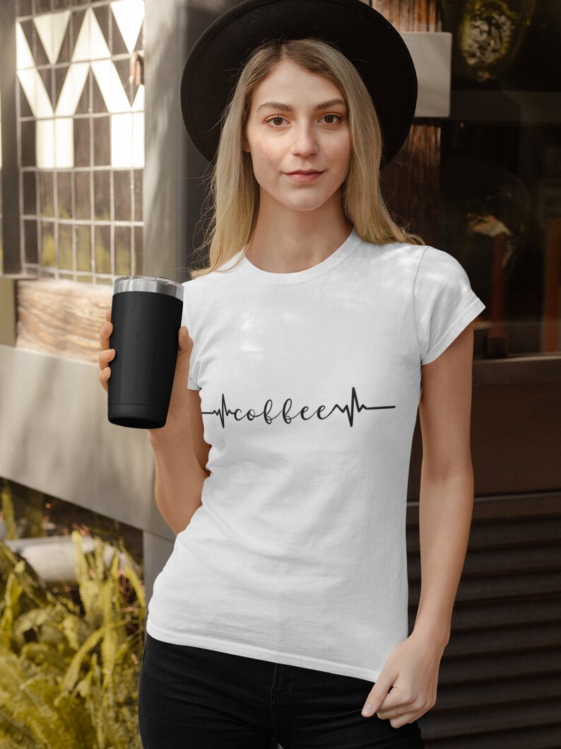 May include: A white t-shirt with a black heartbeat graphic that says "coffee" inside the heartbeat. A person is wearing the shirt and holding a black travel mug.