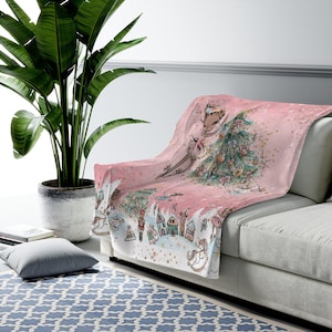 May include: Pink fleece throw blanket with a whimsical Christmas scene featuring a ballerina, a Christmas tree, and a nutcracker. The background is a light pink with gold glitter.