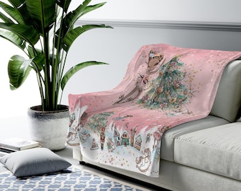 May include: Pink fleece throw blanket with a whimsical Christmas scene featuring a ballerina, a Christmas tree, and a nutcracker. The background is a light pink with gold glitter.
