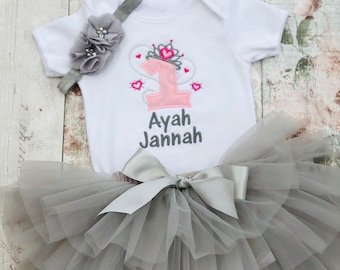 May include: A gray tulle tutu with a silver satin bow, a white onesie with a pink number one, a crown, and hearts, and a gray headband with a flower. The onesie is personalized with the name "Ayah Jannah".