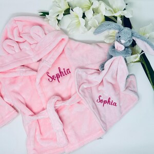 Könnte beinhalten: Rosa Kapuzenbademantel mit Hasenohren und eine passende rosa Sicherheitsdecke mit einem grauen Hasen. Beide Artikel sind mit dem Namen "Sophia" bestickt.