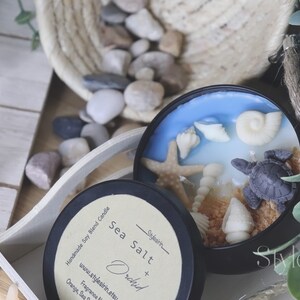 May include: A black candle jar with a blue and white wax design featuring seashells, a starfish, and a turtle. The jar has a label that reads "Handmade Soy Blend Candle Sea Salt + Orchid" and "www.stylesirin.etsy.com".