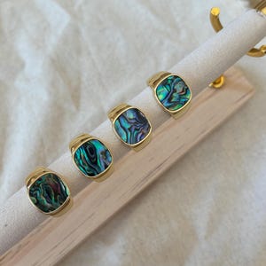 May include: Four gold rings, each featuring a rectangular abalone shell inlay. The shells have iridescent green, blue, and purple hues. The rings are displayed on a light-coloured, fabric-covered jewellery stand.
