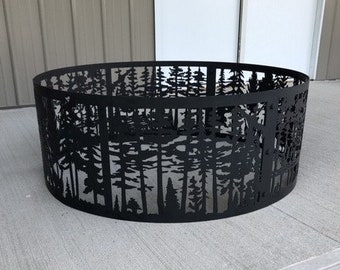 May include: Black metal fire pit ring with a cut-out design of trees. The ring is sitting on a gray concrete surface.