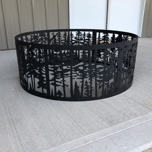 May include: Black metal fire pit ring with a cut-out design of trees. The ring is sitting on a gray concrete surface.