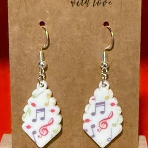 May include: A pair of white, teardrop-shaped earrings with a musical note design. The earrings feature a silver hook and are decorated with colorful musical notes and treble clefs. The background card reads "Handmade with love."
