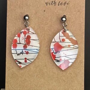 May include: A pair of handmade earrings with a white background and red and black abstract designs. The earrings are made of clear acrylic and have silver hooks.