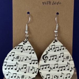 May include: A pair of teardrop-shaped earrings with a white background and black musical notes. The earrings are made of wood and have silver hooks. The earrings are handmade with love.