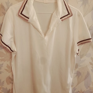 May include: White short-sleeved shirt with a V-neck collar. The collar and sleeve cuffs feature brown and pink stripes. The shirt is hanging on a white hanger against a patterned background.