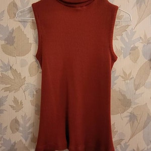 May include: A sleeveless turtleneck sweater in a rust orange color. The sweater is made of a ribbed knit fabric.