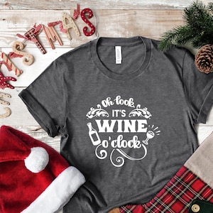 May include: A gray t-shirt with the text "Oh look, it's wine o'clock" in white. The shirt is decorated with a wine bottle and a wine glass graphic. The shirt is on a white wooden background with a red Santa hat, a plaid stocking, and a small gift box.