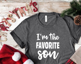 May include: A gray t-shirt with the text "I'm the favorite son" in white lettering. The shirt is on a white wooden background with a red and white stocking, a red Santa hat, and a wooden Christmas sign.
