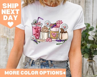 Pode incluir: Camiseta branca com um gráfico de quatro bebidas de café diferentes com flores e um coração rosa em cada bebida. O texto "Happy" está em uma das xícaras de café.