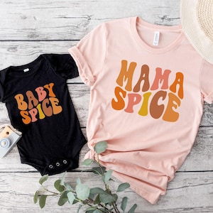 May include: A black baby bodysuit and a light pink t-shirt. The bodysuit has the text "BABY SPICE" in orange and yellow. The t-shirt has the text "MAMA SPICE" in orange, pink, and yellow. A straw hat is visible in the background.