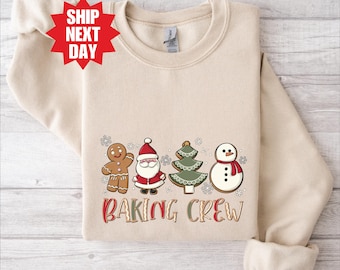 Pode incluir: Camisola bege com um design festivo "Baking Crew". O gráfico inclui personagens de homem de gengibre, Papai Noel, árvore de Natal e boneco de neve. A camisola tem gola redonda e mangas compridas.