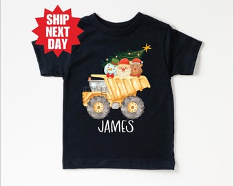 Może przedstawiać: Czarny t-shirt z żółtą wywrotką przewożącą choinkę, bałwana, Świętego Mikołaja i niedźwiedzia. Imię "JAMES" jest wydrukowane na biało. Czerwony graficzny wybuch gwiazdy z napisem "SHIP NEXT DAY" znajduje się po lewej stronie u góry.