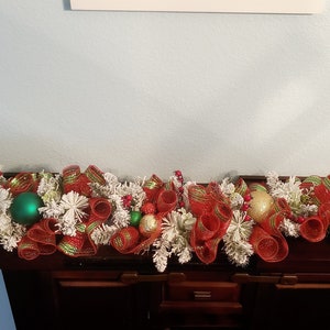 May include: A red and green Christmas garland with white faux snow, red and green ribbon, and gold and green baubles.