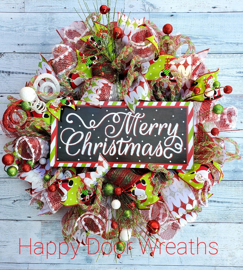 May include: A festive Christmas wreath with a black sign that reads "Merry Christmas" in white script. The wreath features red, green, and white mesh, Santa Claus ribbons, and decorative ornaments. The wreath is on a light blue wooden background.