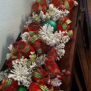 May include: A red and green Christmas garland with white faux snow, green and gold baubles, and red and green ribbon bows.