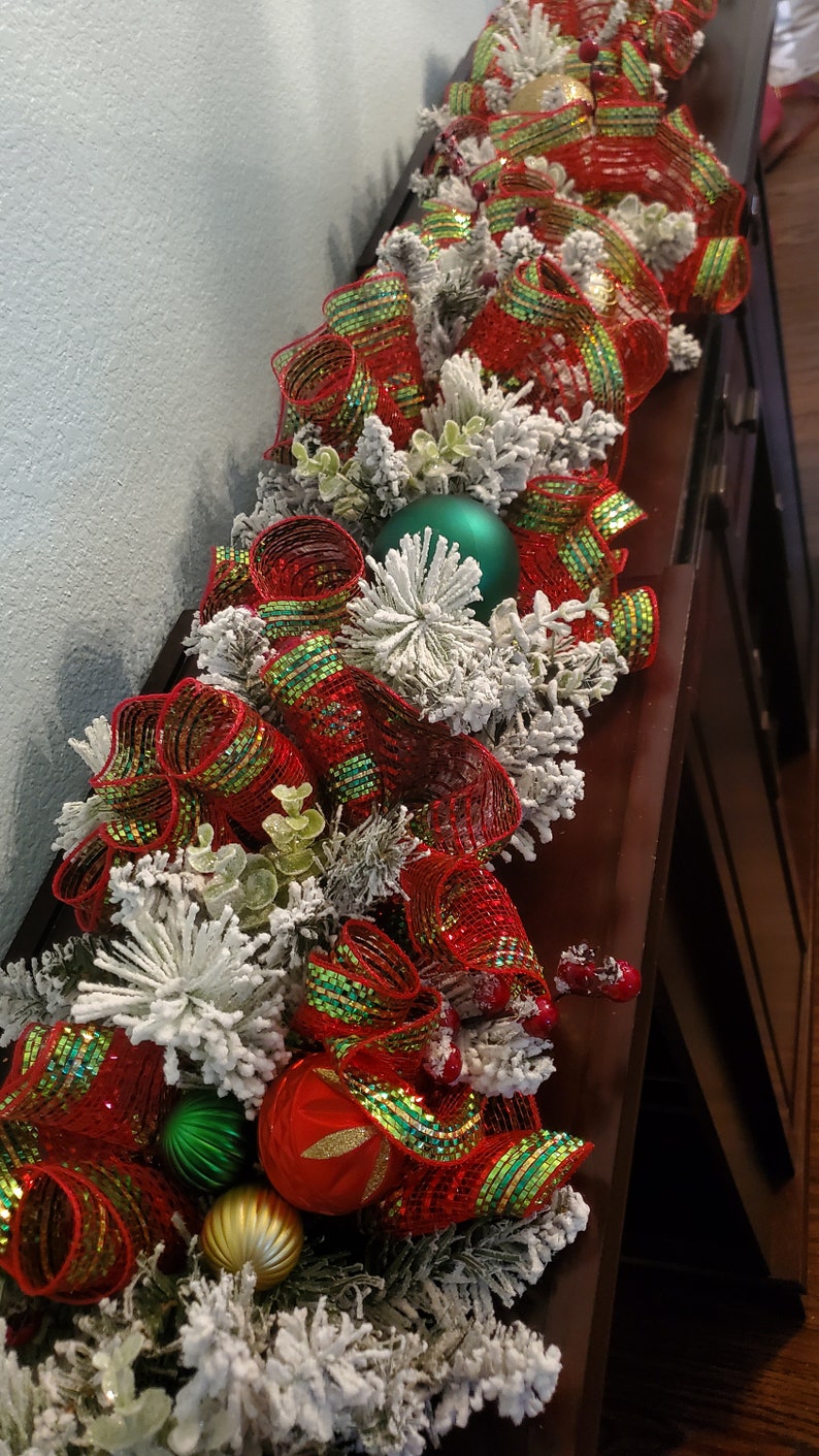 May include: A red and green Christmas garland with white faux snow, green and gold baubles, and red and green ribbon bows.