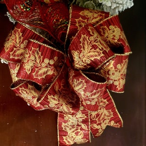 May include: A red and gold Christmas bow with a holly leaf pattern. The bow is made of a shiny, textured fabric.