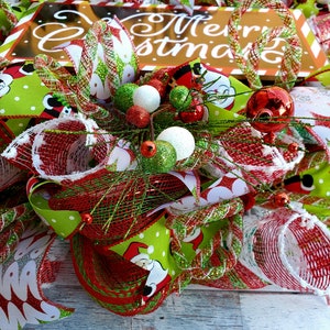 May include: A festive Christmas wreath with a sign that says "Merry Christmas". The wreath features red, green, and white ribbons, ornaments, and mesh. Santa Claus is featured on the green ribbon.