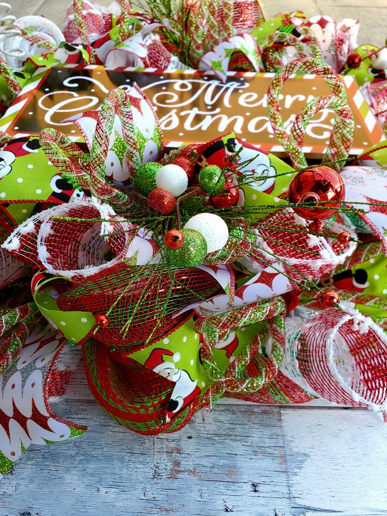 May include: A festive Christmas wreath with a sign that says "Merry Christmas". The wreath features red, green, and white ribbons, ornaments, and mesh. Santa Claus is featured on the green ribbon.