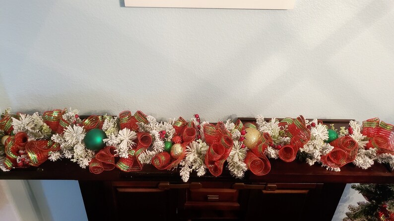 May include: A red and green Christmas garland with white faux snow, red and green ribbon, and gold and green baubles.