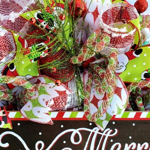 May include: A festive Christmas wreath with red, green, and white accents. The wreath features decorative ribbons, ornaments, and a sign that reads "Merry Christmas" in white script. The wreath is adorned with red and green accents.