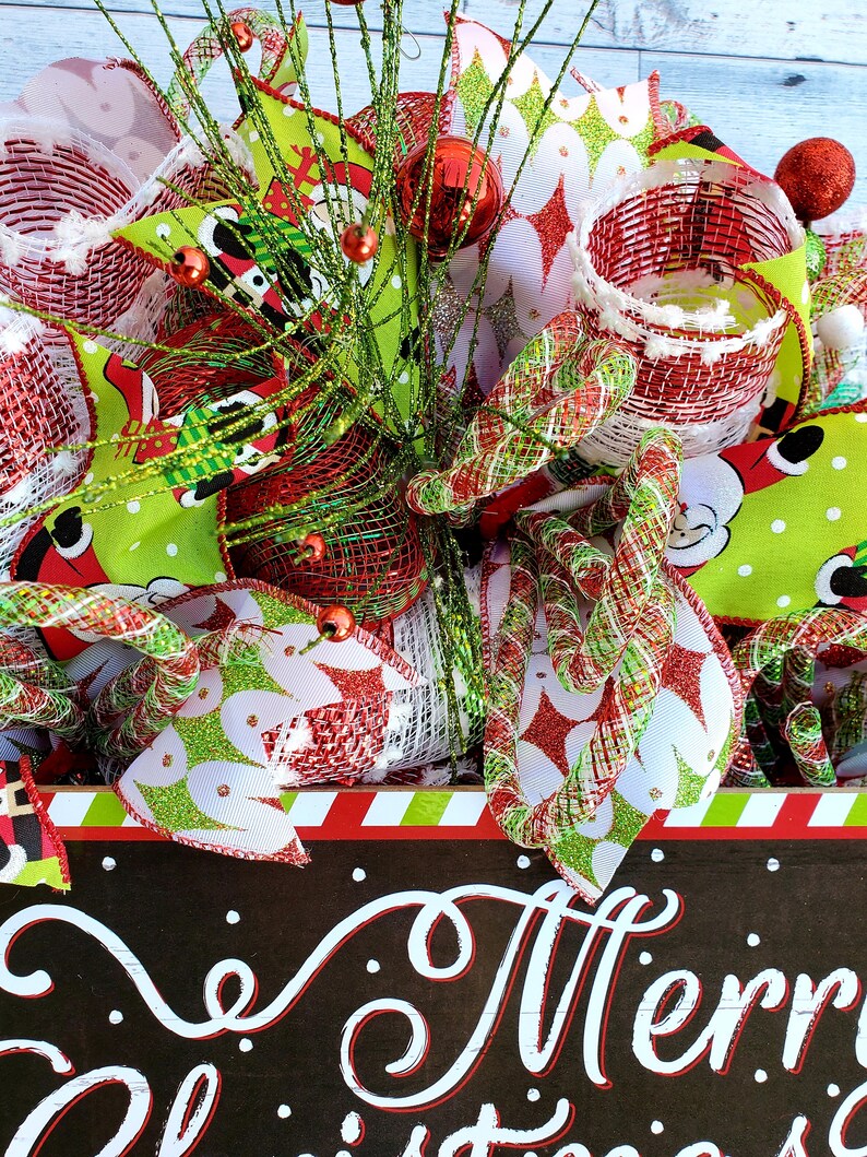 May include: A festive Christmas wreath with red, green, and white accents. The wreath features decorative ribbons, ornaments, and a sign that reads "Merry Christmas" in white script. The wreath is adorned with red and green accents.
