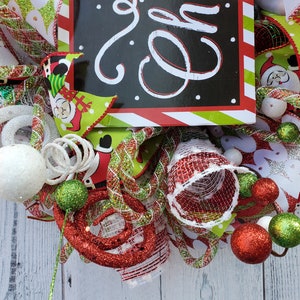 May include: A festive Christmas wreath with a black sign that says "Joy" in white script. The wreath features red, green, and white ribbons, Santa Claus accents, and decorative ornaments. The wreath is adorned with glittery balls and swirls.