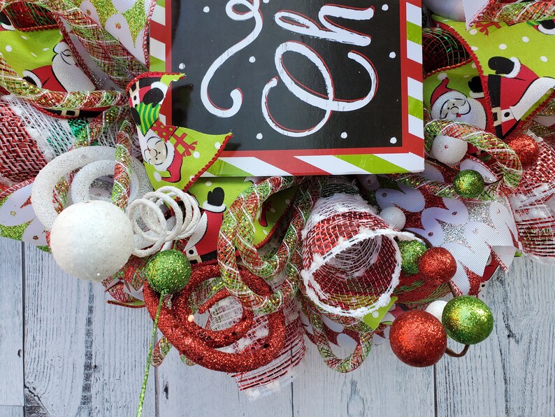 May include: A festive Christmas wreath with a black sign that says "Joy" in white script. The wreath features red, green, and white ribbons, Santa Claus accents, and decorative ornaments. The wreath is adorned with glittery balls and swirls.