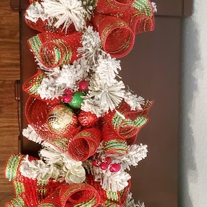 May include: A Christmas garland featuring red and green ribbon, white faux snow, and gold and green baubles.