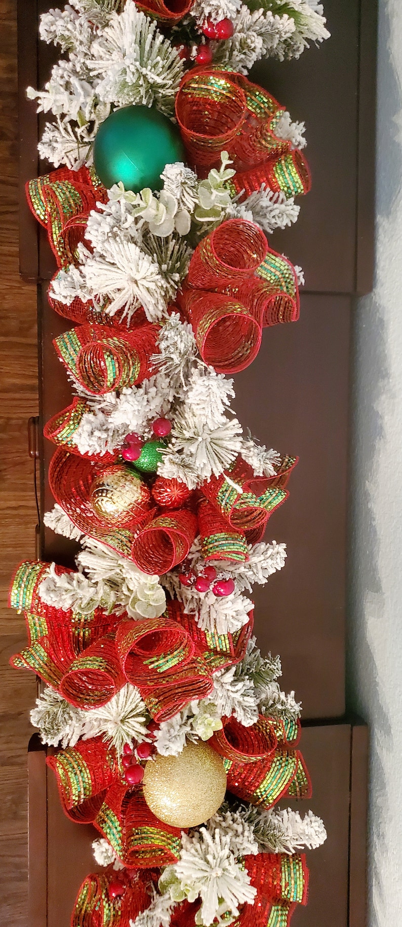 May include: A Christmas garland featuring red and green ribbon, white faux snow, and gold and green baubles.