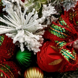 May include: A close-up of a Christmas wreath featuring red and green ribbon, white flocked pine branches, and gold and green baubles.