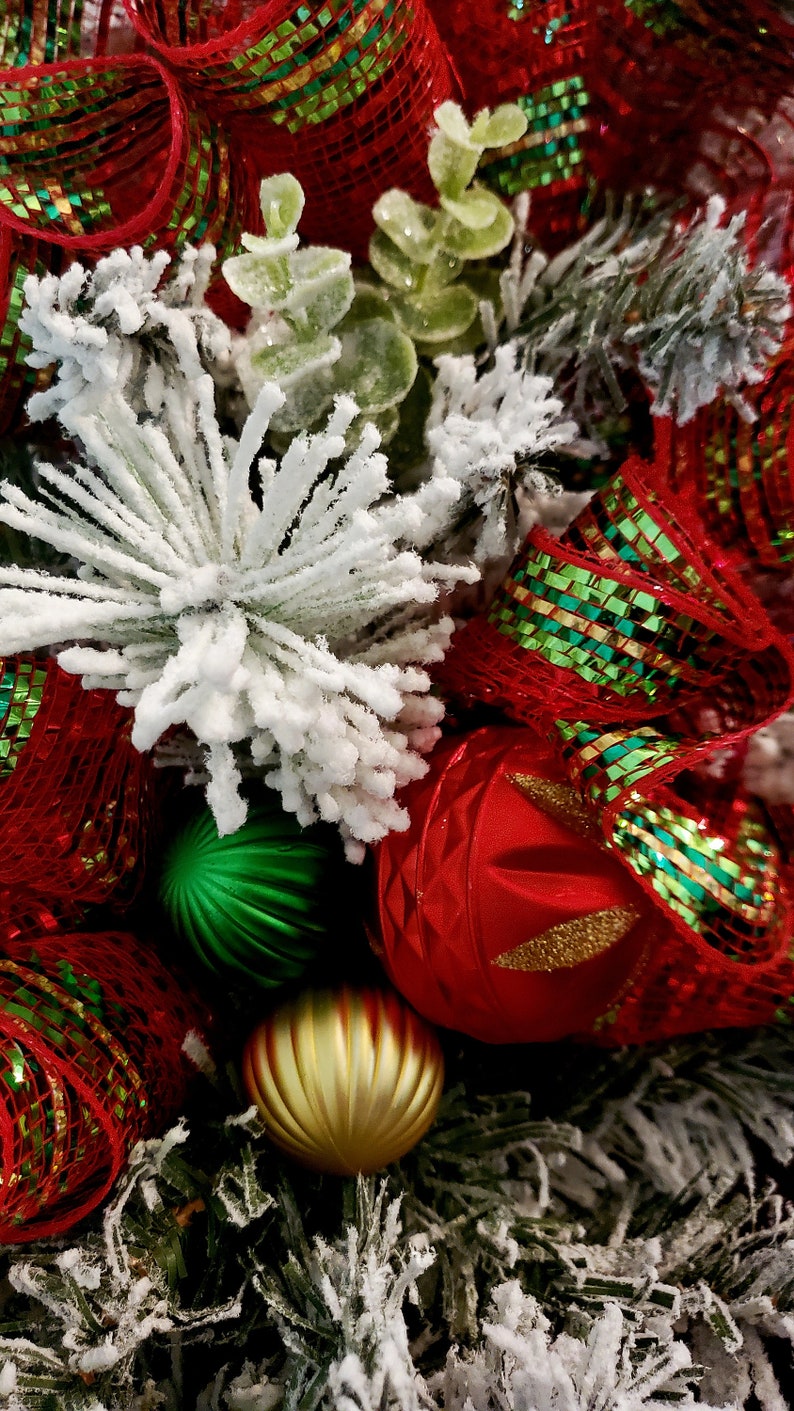 May include: A close-up of a Christmas wreath featuring red and green ribbon, white flocked pine branches, and gold and green baubles.
